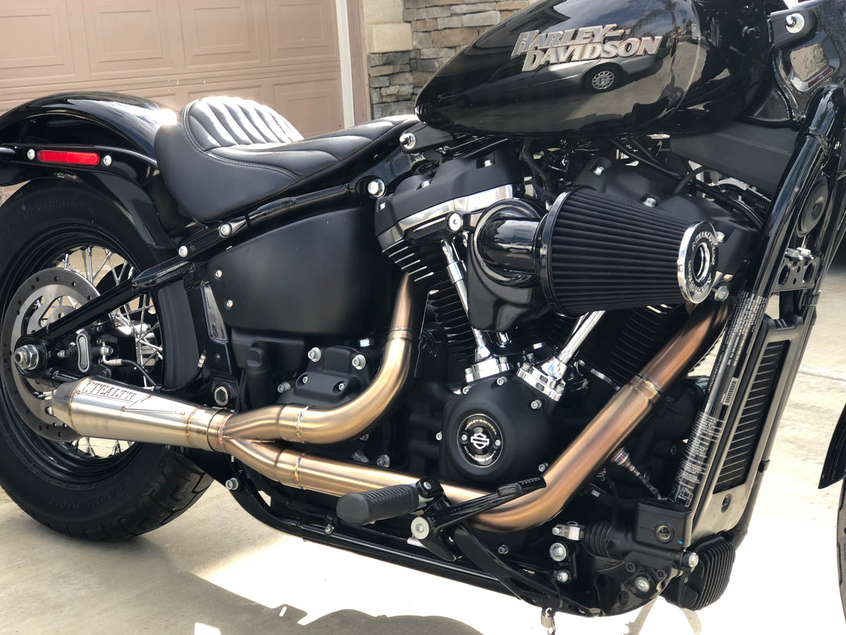A black motorcycle with the focus on the exhaust system, which is silver in color. The motorcycle is parked against a plain wall backdrop., photo 1 of 36